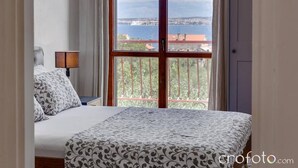 A bedroom with a bed, a nightstand, a lamp, and a window with a view of a balcony and a body of water. - 3-bedroom Holiday House Giovanni (Preko)