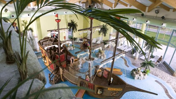 Un modelo de barco en una fuente de agua con ambiente tropical, incluyendo palmeras y una zona de arena con sillones de descanso.