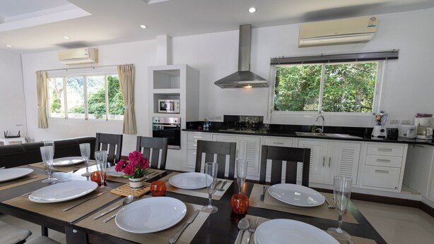 A modern kitchen with white cabinets, a large window, and a dining area set for a meal. - Phikun Private Pool Villa (Chalong)