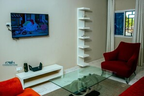 A modern living room with a flat-screen TV, a glass coffee table, a red armchair, and a white shelving unit.