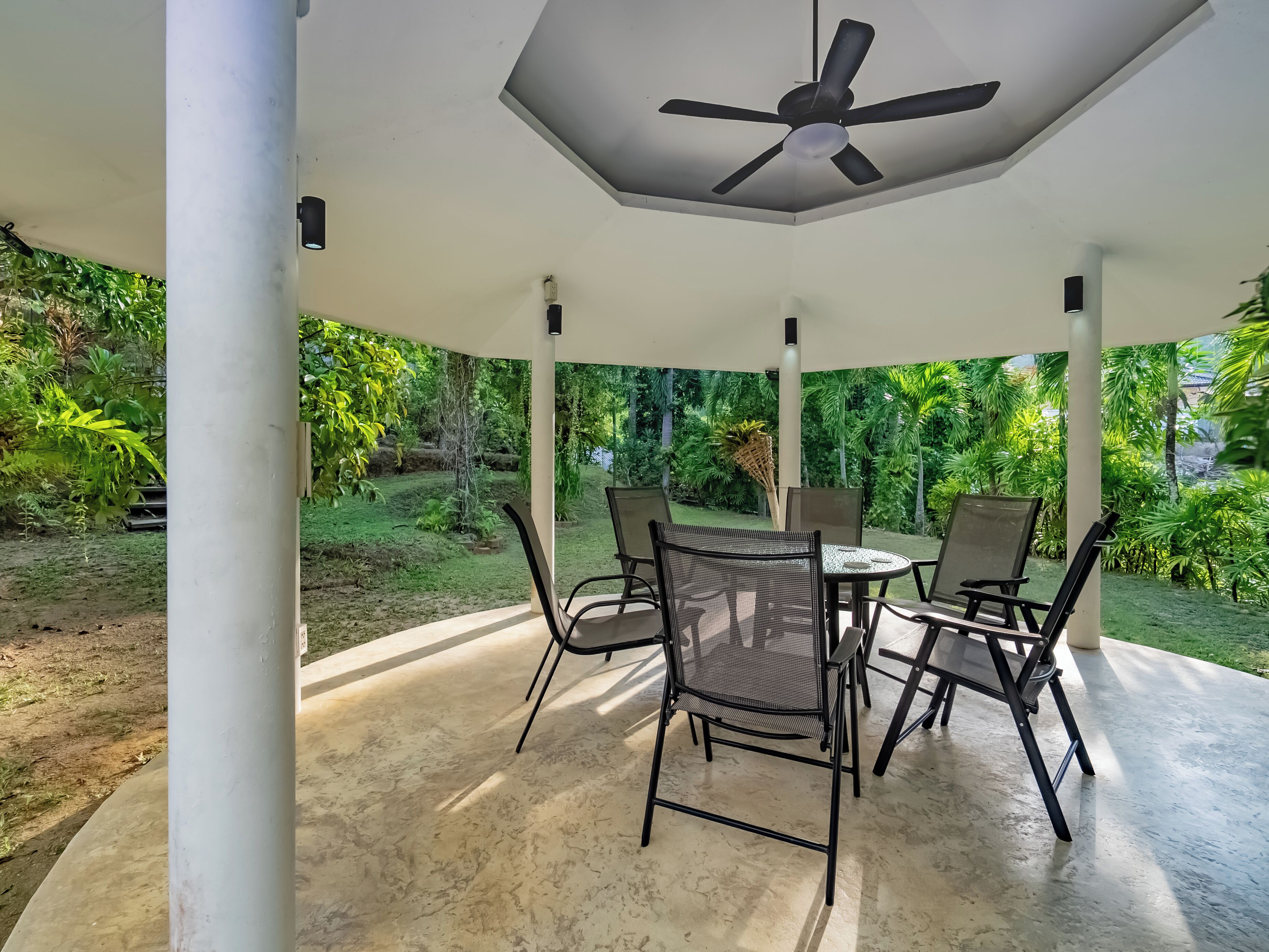 A covered outdoor seating area with a ceiling fan, surrounded by lush greenery.