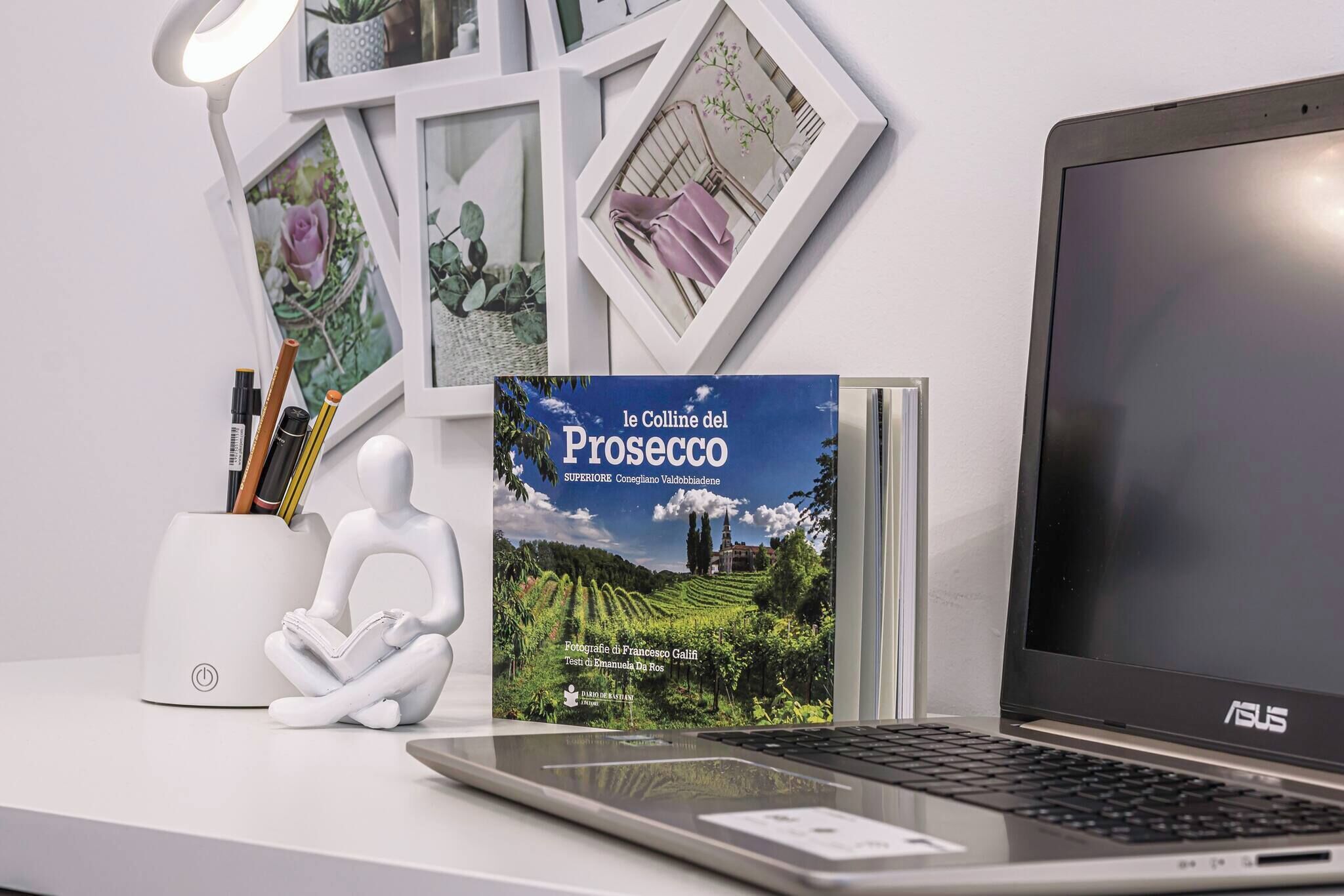 Een laptop op een bureau met een boek getiteld 'le Colline del Prosecco' en een witte lamp.