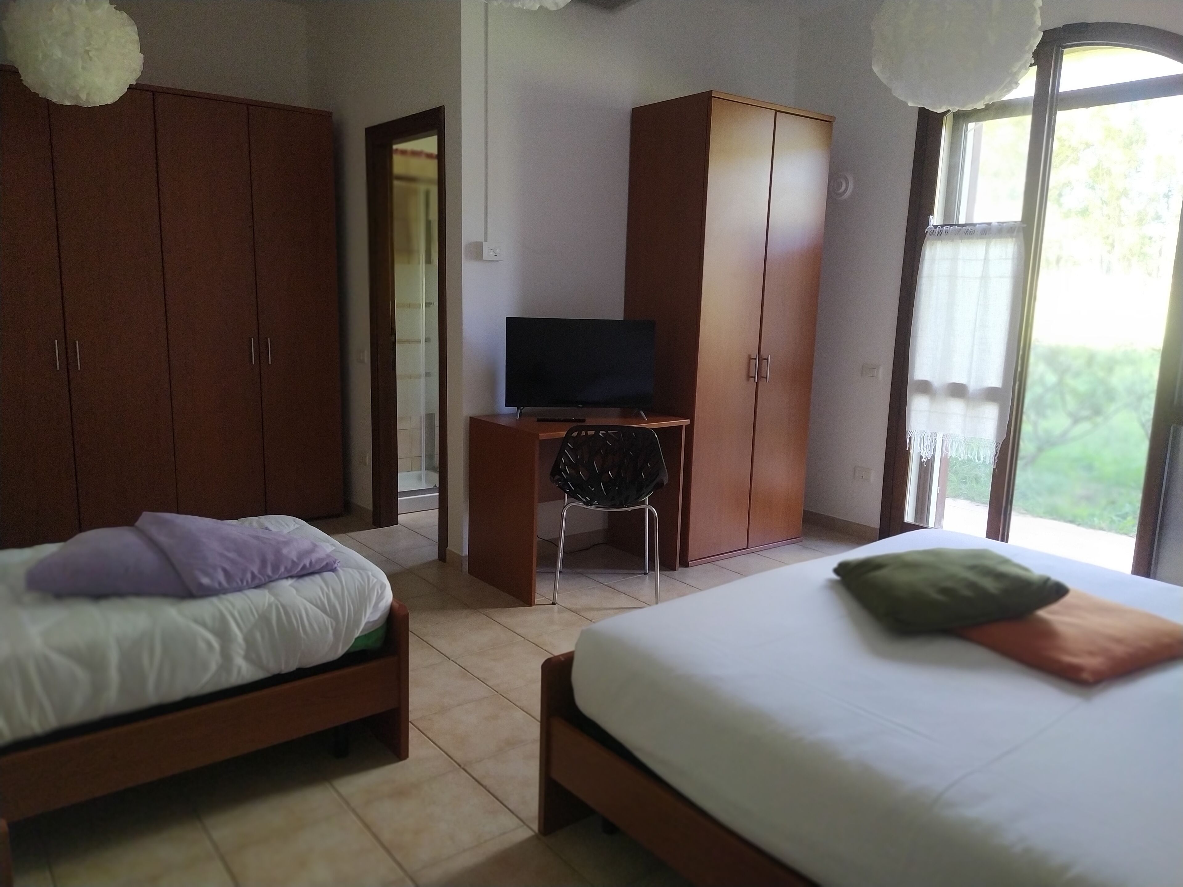 A bedroom with two beds, a desk with a TV, wooden wardrobes, and a window with a view.