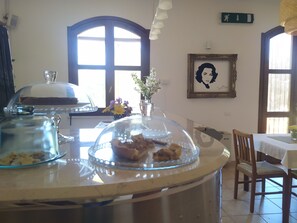 A dining area with a round table, chairs, a glass display case with food, a window with a view, and a framed picture on the wall. - Room in B&B - "note di Stelle" Resort in Marrubiu (Marrubiu)