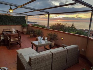 Une terrasse avec une table, des chaises et un canapé, d’où l’on peut admirer le coucher de soleil sur la mer.