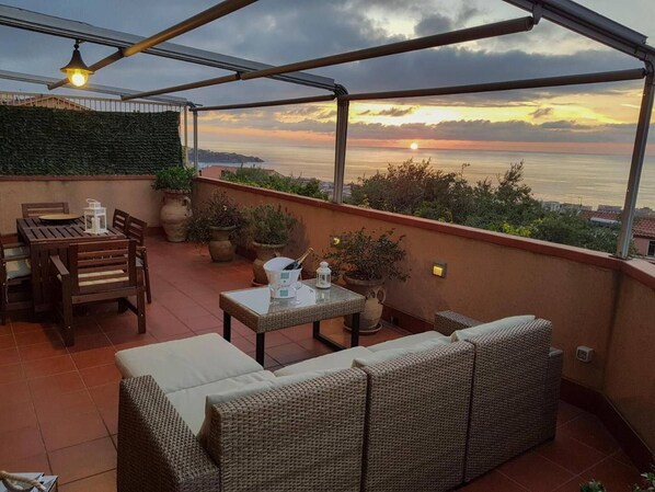 A terrace with a table, chairs, and a sofa, overlooking a sunset over the sea.