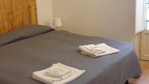 Een slaapkamer met een bed, twee handdoeken gevouwen op een plankje, een houten hoofdeinde, een bedlampje en een raam.