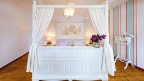 Una cama con dosel blanco y cortinas, una mesita de noche con lámpara y una obra de arte colgada en la pared.