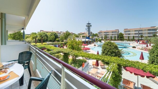 A balcony with a view of a swimming pool, lounge chairs, and a tower in the background. - Super Villaggio Planetarium Resort 1 Bedroom Apartment Sleeps 4 (San Michele al Tagliamento)
