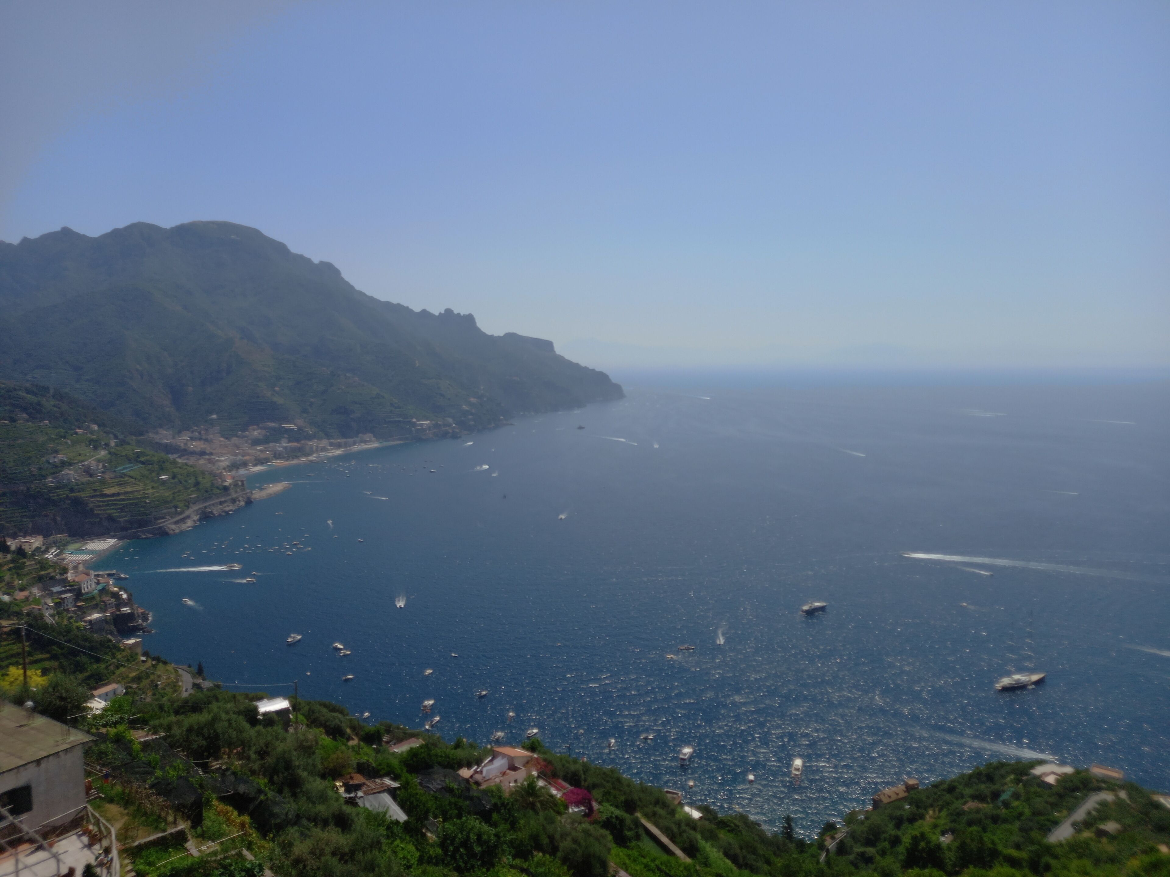 Vista costera con barcos en el agua, montañas al fondo y un pequeño poblado con edificios.