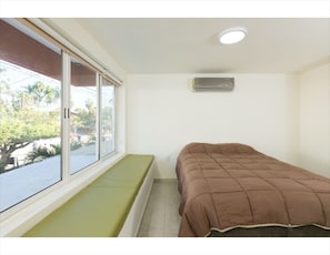 Quarto com cama, janela com vista para palmeiras, luminária no teto e aparelho de ar condicionado.