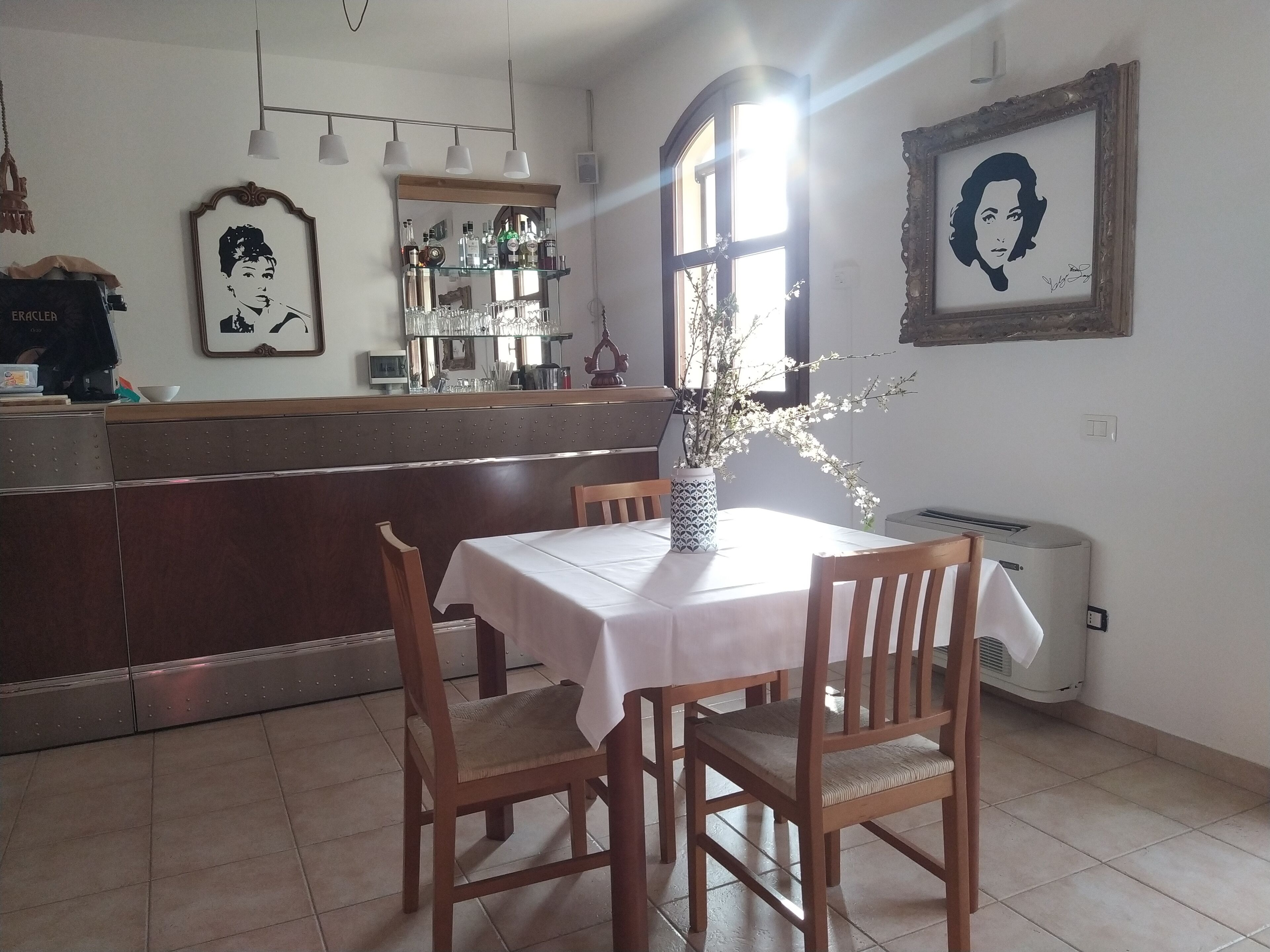 A dining area with a table set for two, wooden chairs, a vase with branches, and a bar area in the background.