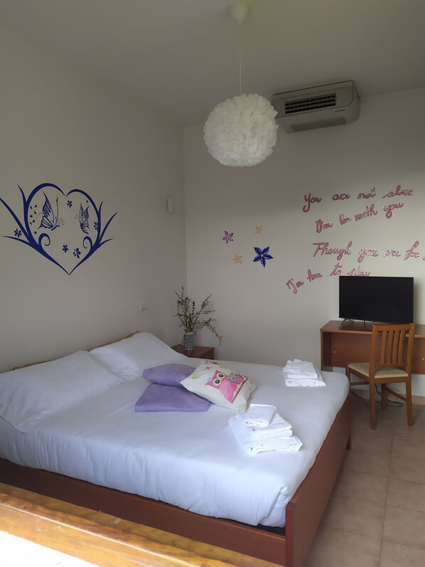 Cama bem arrumada com almofadas brancas e roxas, uma pequena escrivaninha com cadeira, uma televisão e adesivos decorativos na parede.