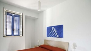 A bedroom with a bed, a nightstand, a lamp, a window with blinds, and a blue abstract painting on the wall. - Dolce Mare 1 - Ground Floor With Garden (Modica)