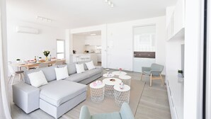 A modern living room with a grey sectional sofa, a dining area with a wooden table, and a kitchen in the background. - Protaras Olivine Villa Ol9 (Paralimni)