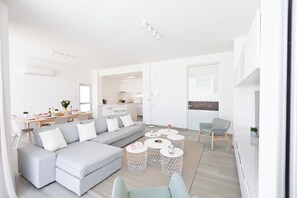 A modern living room with a grey sectional sofa, a dining area with a wooden table, and a kitchen in the background. - Protaras Olivine Villa Ol9 (Paralimni)
