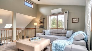 Una sala de estar moderna con un sofá color beige, un reposapiés a juego y un plaid azul tejido. Se ve una escalera con barandilla de madera, una ventana con cortinas y un equipo de aire acondicionado en la pared.