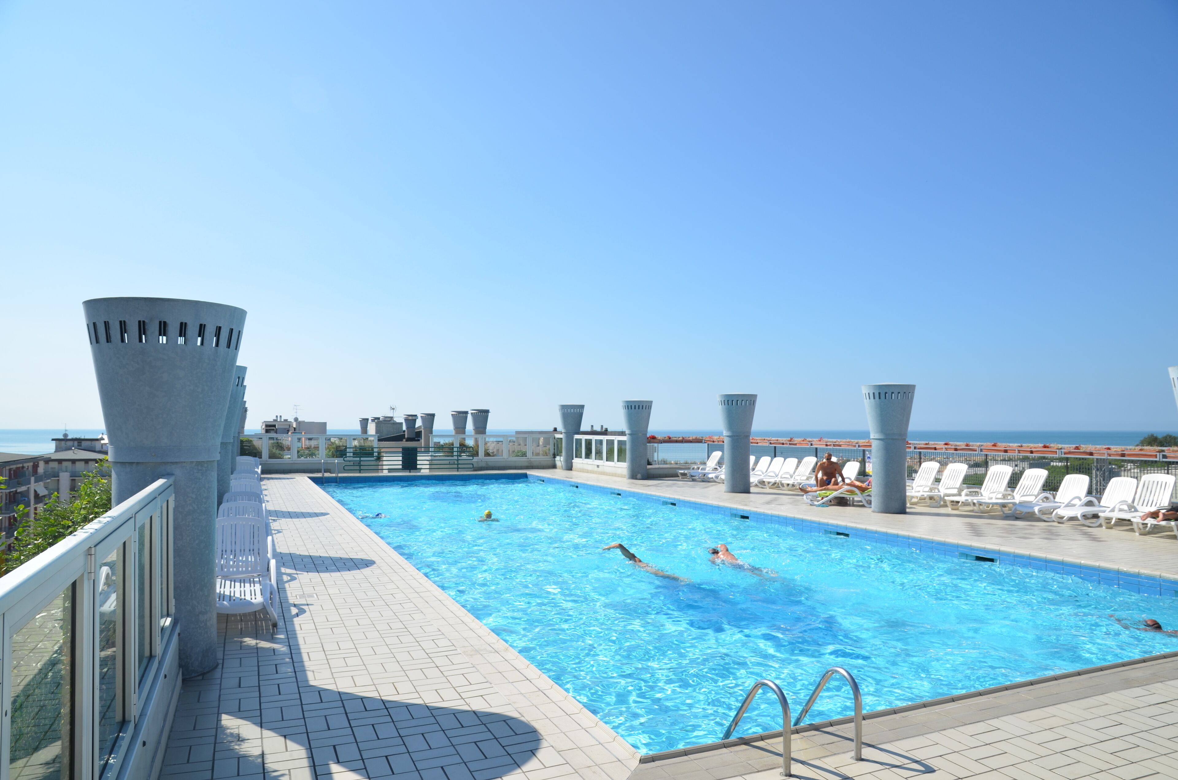 Una piscina en la azotea con agua azul cristalina, rodeada de sillones blancos y vistas al mar.