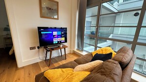 A living room with a large window, a flat-screen TV, and a brown sofa with a yellow throw blanket. - Modern & Stylish 1 Bedroom Flat (London)