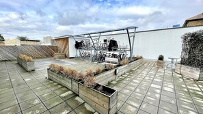 Terraço no telhado com piso de azulejos, vasos de madeira com plantas, suporte de bicicleta de metal e um banco.