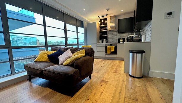 A modern living room with a brown sofa, large windows, and a kitchen area with a black TV and white cabinets. - Modern & Stylish 1 Bedroom Flat (London)