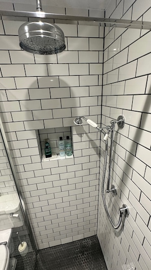 Une salle de bain avec des murs recouverts de carreaux de céramique blancs rectangulaires, une douche équipée d’un pommeau de douche amovible et une petite étagère contenant des articles de toilette.