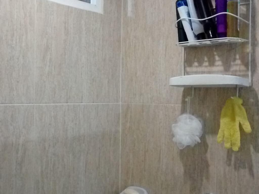 Een badkamer met een betegelde muur, een wit plankje met toiletartikelen, en een gele handschoen aan de muur.