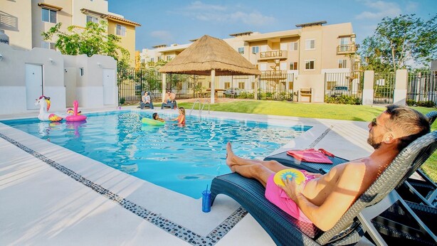 A swimming pool with lounge chairs, a thatched-roof structure, and modern buildings in the background. - Mayakoba Cozy 2-bedroom Apartment With Amenities (Playa del Carmen)