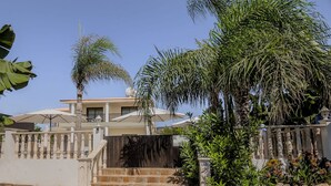 Una casa a due piani con un balcone, circondata da palme e un recinto bianco.