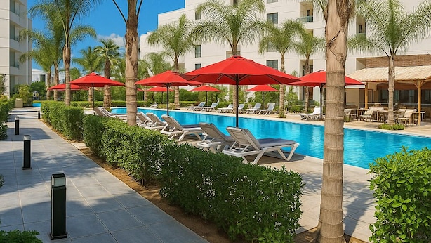 A pool area with lounge chairs and red umbrellas, surrounded by palm trees and a modern building. - Port Said City, Damietta Port Said Coastal Road Num3070 (Al-Manasrah)
