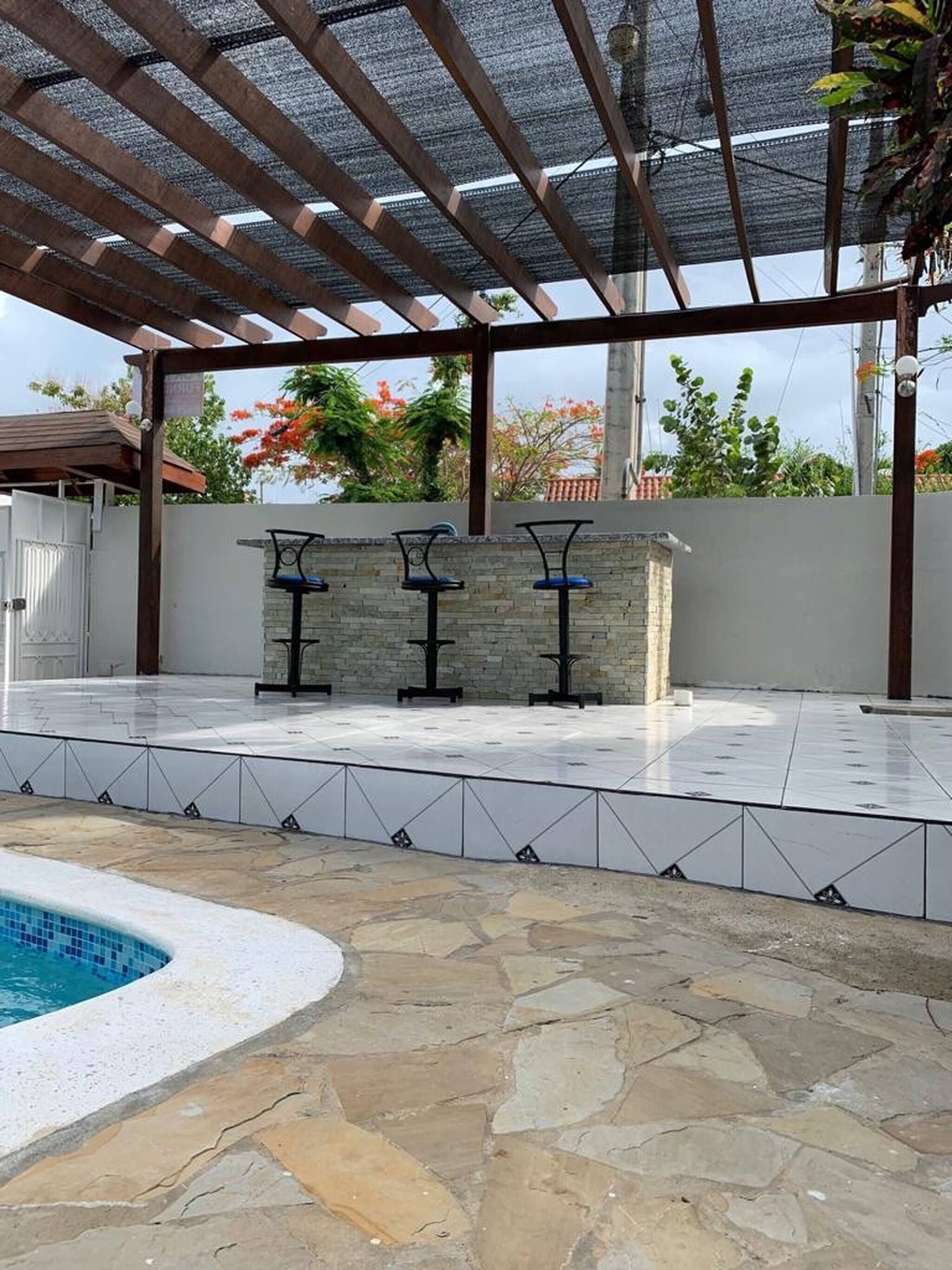 Un espace extérieur aménagé en terrasse, avec une piscine, un sol en pierre et trois tabourets de bar noirs adossés à un mur de pierre.