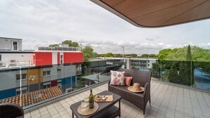 A balcony with wicker furniture, a bottle, and glasses, overlooking a cityscape with buildings and greenery. - Elegant Residenza Monica 2 Two Bedroom C6 Sleeps 6 (Lignano Sabbiadoro)