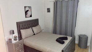 Un dormitorio con cama, mesita de noche, lámpara, un cuadro en la pared y cortina.
