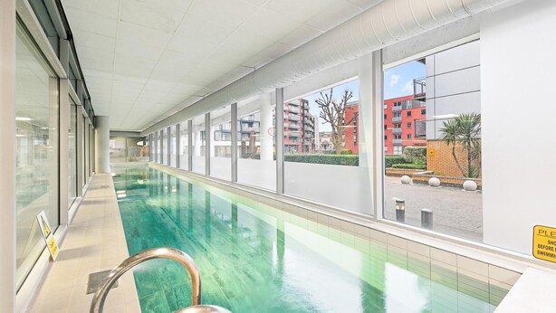 Una piscina interior larga con paredes de vidrio transparente, una barandilla de metal y vistas a un edificio rojo en el exterior.