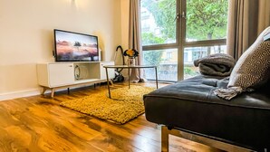 Una sala de estar moderna con suelo de madera, un sofá de cuero negro, un mueble blanco y un televisor que muestra una escena de la naturaleza.