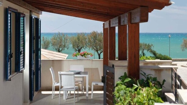 A balcony with a wooden pergola, white chairs, and a table. It overlooks a coastal area with trees and a view of the sea. - Dolce Mare 4 - Sea Front - Large Balcony - Sandy Beach (Modica)