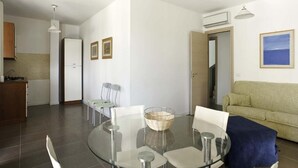 A modern living room with a glass dining table, white chairs, a blue sofa, and a kitchen area in the background. - Dolce Mare 4 - Sea Front - Large Balcony - Sandy Beach (Modica)