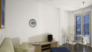 A modern living room with a sofa, a small TV on a wooden stand, a dining table with chairs, and a large window with blinds. - Dolce Mare 4 - Sea Front - Large Balcony - Sandy Beach (Modica)