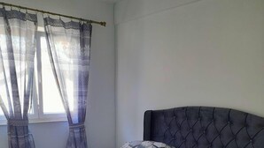 Una cama bien hecha con ropa de cama estampada, un cabecero tapizado oscuro y una ventana con cortinas de gasa.
