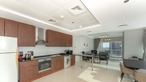 A modern kitchen with wooden cabinets, a built-in oven, and a washing machine. There is a dining area with a table and chairs, and a large window offering a city view. - Homesgetaway - JLT Lake Views 1BR (Dubai)