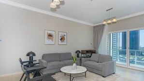 A modern living room with a grey sofa, a round coffee table, and a dining area with a black table and chairs. There are two framed artworks on the wall, and a city view through large windows. - Homesgetaway - JLT Lake Views 1BR (Dubai)