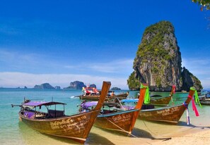 A beach with several traditional wooden boats docked, a clear blue sky, and a prominent rocky island in the background. - Centre Point Patong (Patong)