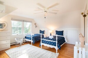 A bedroom with two beds, a ceiling fan, a dresser, a rocking chair, and a window with a view of greenery.