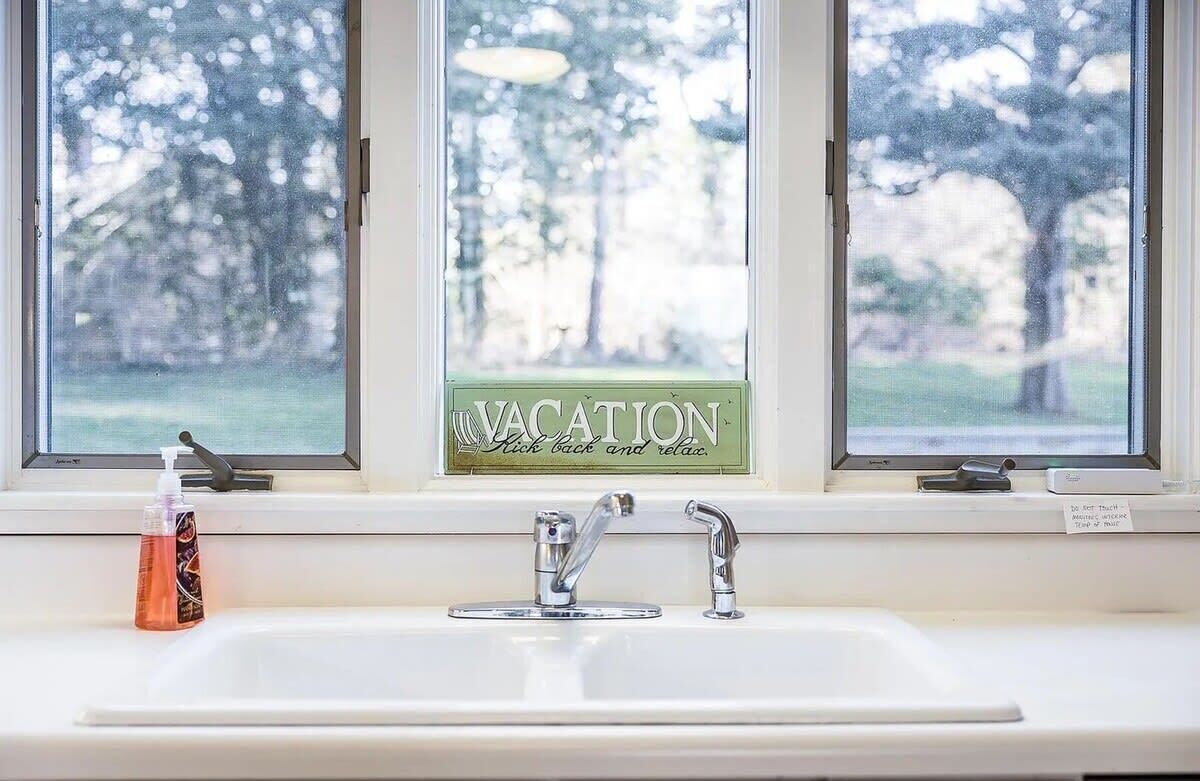 Ein Badezimmer mit Waschbecken, zwei Wasserhähnen und einem Fenster mit der Aufschrift 'VACATION'.