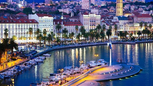 Un porto turistico con barche ormeggiate, una passeggiata sul lungomare e edifici storici sullo sfondo.