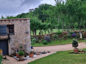 Un bâtiment rustique en pierre, avec un chemin de gravier menant à un jardin où poussent différentes plantes et un petit arbre.