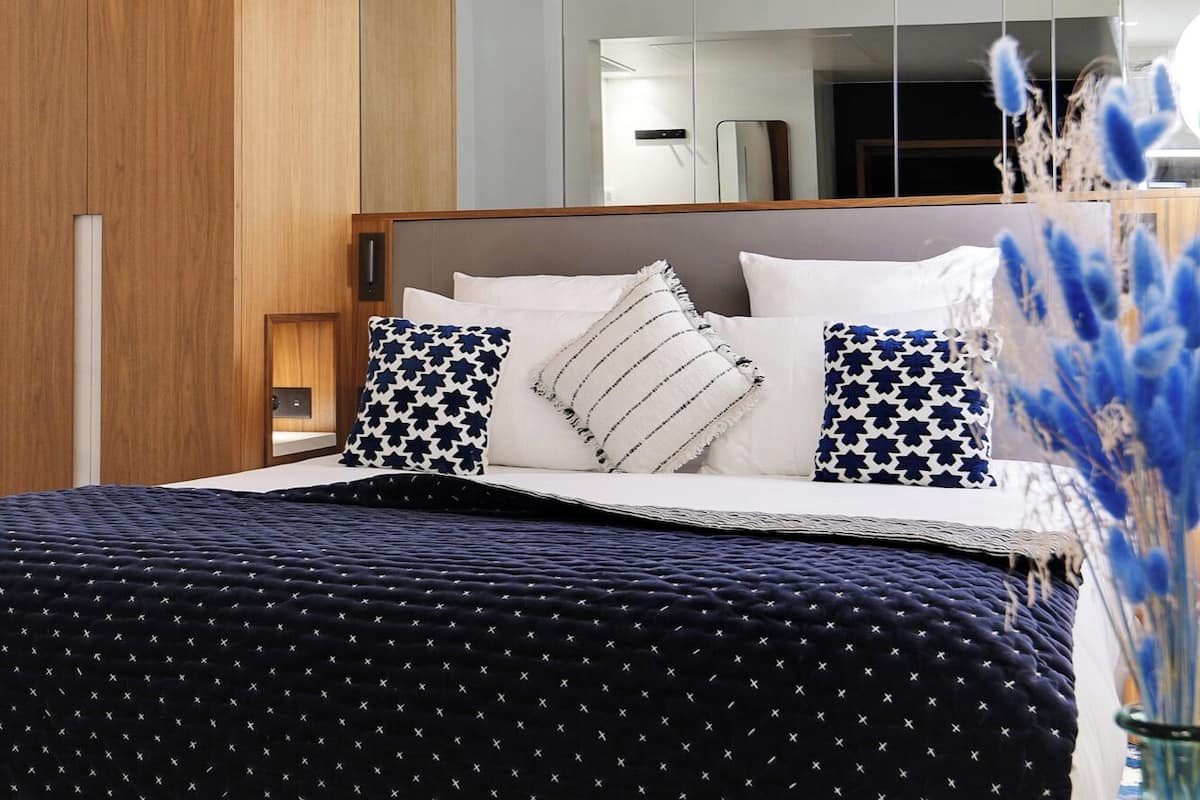 A bedroom with a bed, pillows, a nightstand, a mirror, and a shelf with books and decorative items.
