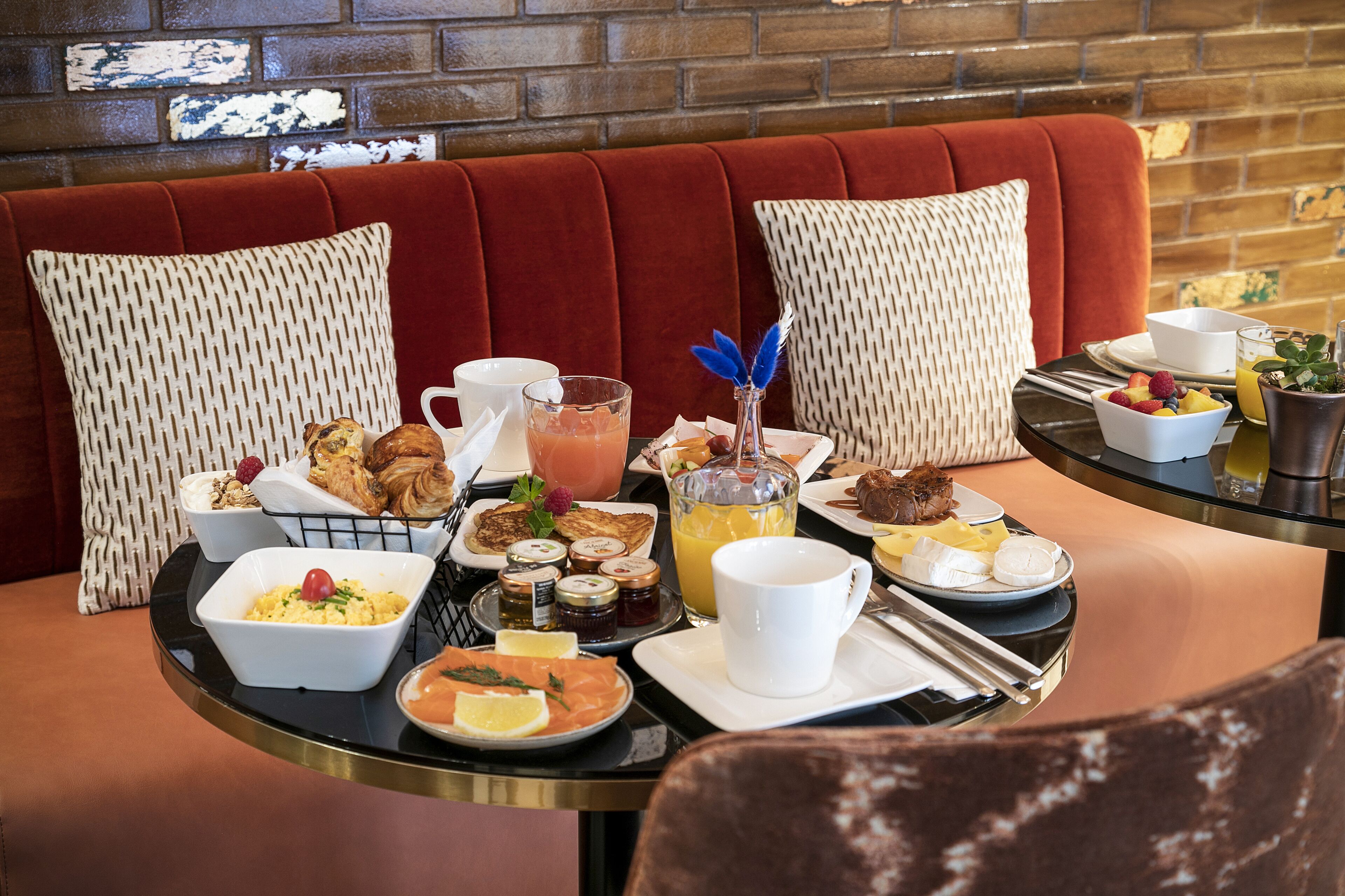 A breakfast buffet with various dishes and drinks on a table, set against a brick wall and red upholstered seating.