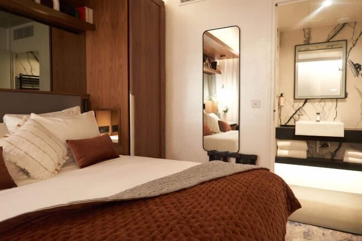 A bedroom with a bed, a large mirror, a bedside table, and a bathroom visible through an open door.