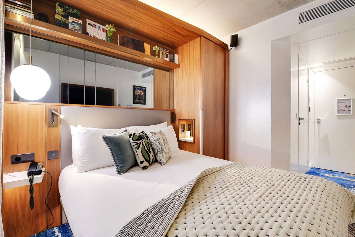 A neatly arranged hotel room with a large bed, a bedside table with a phone, a mirror above the bed, and a wooden shelf with various items.
